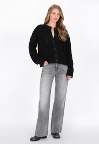 Fekete kötött kardigán kerek nyakkivágással, elülső gombokkal és laza szabással. Viharos szürke, bővülő nadrággal és fekete cipővel párosítva.