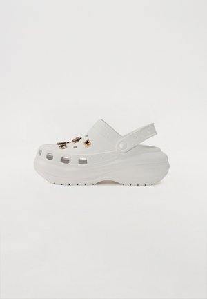 Sabot blanc en matériau léger et durable. Doté de trous de ventilation et orné de quincaillerie décorative. Inclus une sangle arrière.