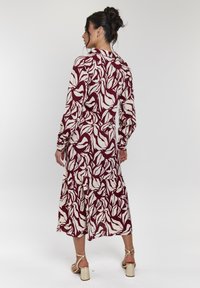 Langes, burgunderfarbenes Kleid mit weißem Blattmuster, mit langen Ärmeln, geraffter Taille und gestuftem Saum. Getragen mit goldenen Sandalen mit Riemchen.