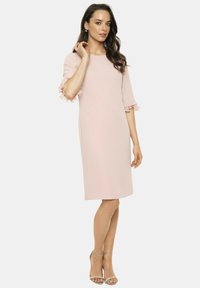 Jasnoróżowa sukienka do kolan z krótkimi falbaniastymi rękawami, prosty krój i gładka tekstura. Dobrana ze srebrnymi sandałami na obcasie.