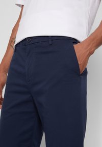 Námořnické modré chinos kalhoty vyrobené z hladkého materiálu, s knoflíkovým zapínáním, bočními kapsami a elegantním designem vhodným pro volnočasové nošení.