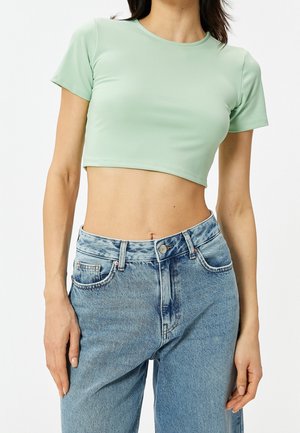 Frau trägt ein hellgrünes, kurzärmeliges, bauchfreies Top und hoch taillierte blaue Denim-Jeans, die Hände entspannt an den Seiten.