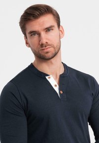 Man med kort brunt hår och skägg som bär en marinblå henley-tröja med vit placket och träknappar, mot en enkel vit bakgrund.