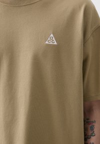 Oliven grøn bomulds T-shirt med rund halsudskæring og korte ærmer, med et lille hvidt trekantet logo på venstre bryst.