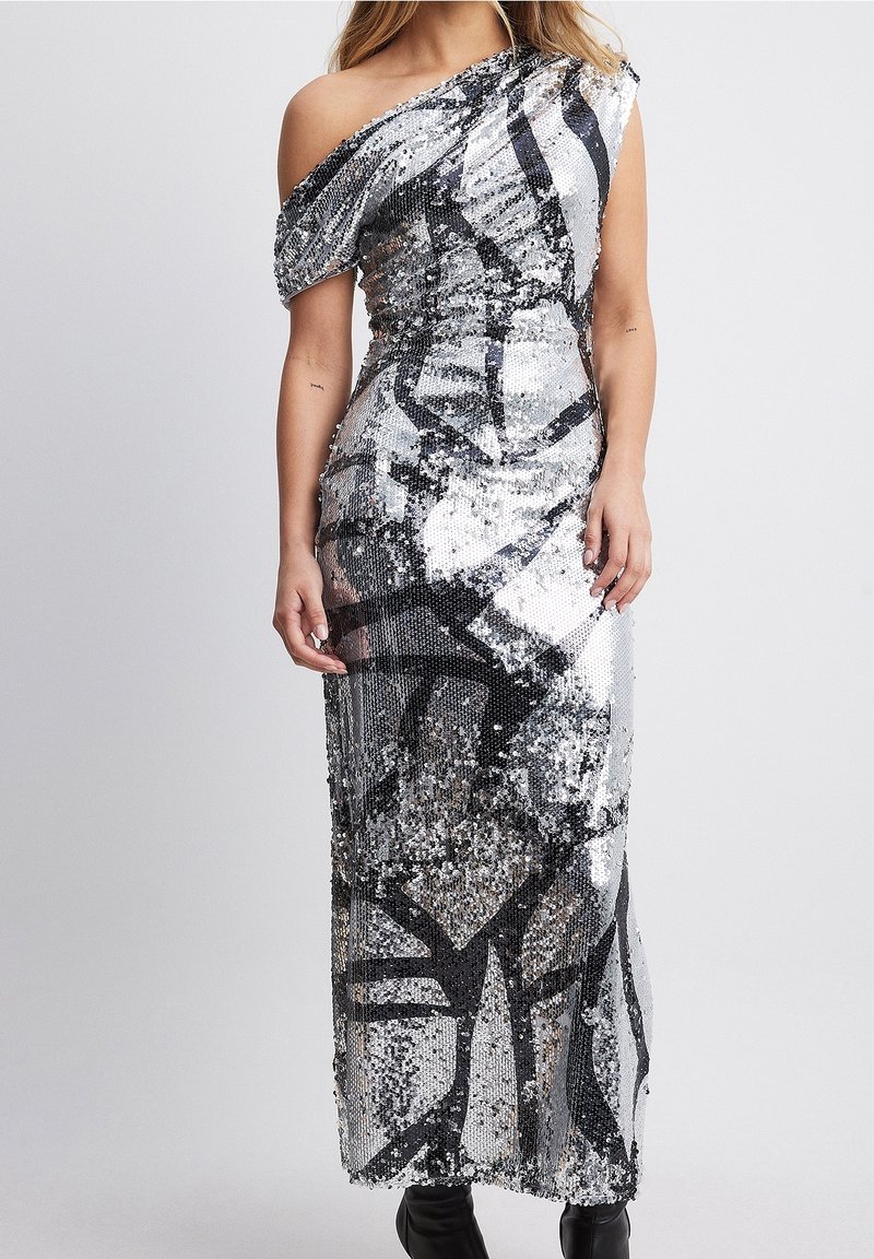 Femme portant une longue robe argentée à sequins à une épaule avec des motifs abstraits noirs, associée à des bottines noires.