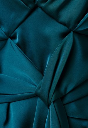 Žalvarinės spalvos audinys su dekoratyvine kilpa ir klostėmis, sukuriantis tekstūruotą, sluoksniuotą raštą.