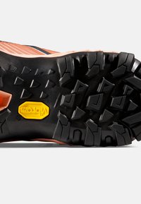 Zwarte rubberen loopzool met diepe profiel, voorzien van een gele Vibram-logo. Bovenkant is een combinatie van perzikkleurige en zwarte materialen.