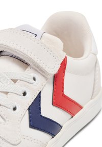 Vit sneaker med ovandel i läder, texturerade röda och blå detaljer, snörning och en kardborreband för extra säkerhet.