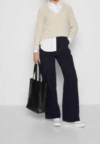 Beige gerippter Pullover über einem weißen Hemd mit Kragen getragen, kombiniert mit dunkelblauen weit geschnittenen Hosen und weißen Plateausneakern. Schwarze Tragetasche.