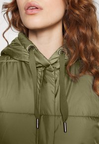 Pikowana kurtka w oliwkowej zieleni z kapturem, z przeszyciami z przodu, metalowymi przelotkami i sznurkami do ściągania z tkaniny. Gładka, błyszcząca faktura.