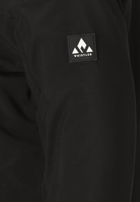 Svart jacka i slätt tyg med en fyrkantig etikett på ärmen med en vit bergslogotyp och ordet "WHISTLER."