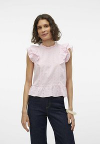 Blouse sans manches rose clair avec des épaules volantes, en tissu texturé, détails œillets et ourlet peplum, associée à un pantalon en denim foncé.