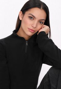 Zwarte fleece pullover met een kwart ritssluiting, zachte textuur en lange mouwen. Model poseert met hand onder de kin, dragend een donkergekleurde spijkerbroek.