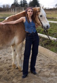 Mlada ženska v traku iz jeansa in kavbojkah stoji ob svetlo rjavem konju v peščenem ograjenem prostoru z lesenimi ograjami in drevesi v ozadju.