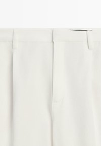 Close-up of white high-waisted trousers with belt loops, visible seam, and hidden front zipper.