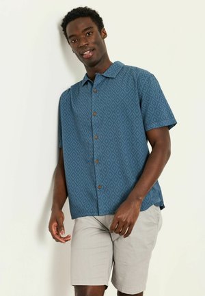 Junger Mann mit einem blau gemusterten kurzärmeligen Hemd und hellbeigen Shorts, stehend vor einer weißen Wand.