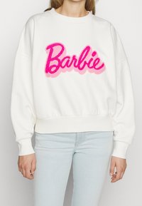 Vit sweatshirt med en rosa, fluffig "Barbie" textlogotyp. Designen har en rund halsringning och ribbade muddar och fåll.