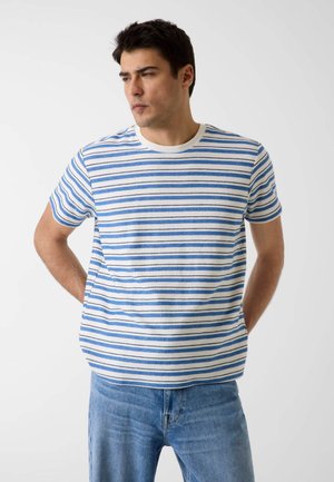 Man die een blauw-wit horizontaal gestreept kortärmelig shirt en blauwe jeans draagt, staat met de handen achter de rug en kijkt naar links.