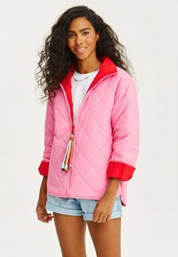 Pletená ružová bunda s červeným golierom a manžetami, so sťahovacou šnúrkou s farebnými akcentmi, nosená cez bielu košeľu a riflové šortky.