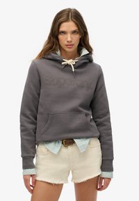 Haut à capuche gris court avec logo "Superdry" en relief, capuche avec cordon de serrage, et poche kangourou, superposé sur une chemise à rayures bleu clair et un short blanc.