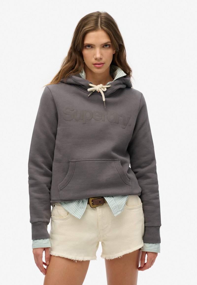 Haut à capuche gris court avec logo "Superdry" en relief, capuche avec cordon de serrage, et poche kangourou, superposé sur une chemise à rayures bleu clair et un short blanc.