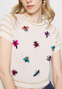 Suéter de punto rosa claro con mangas cortas, que presenta coloridos insectos bordados y un cuello con rayas metálicas doradas.