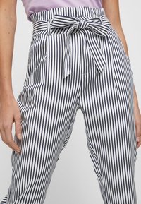 Pantalon rayé blanc et bleu marine, à taille haute, avec un détail nœud papillon et des poches latérales ; le tissu semble léger avec une texture lisse.