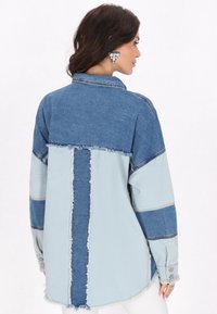 Jeanshemd met een patchworkontwerp in donkerblauwe en lichtblauwe tinten. Heeft franje randen en een knoopdetail op de mouw. Achteraanzicht getoond.