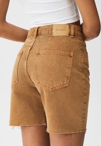 Shorts en denim brun avec un ourlet effiloché, dotés de deux poches arrière et d'un patch en étiquette sur la taille.
