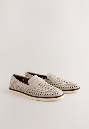 Paire de mocassins beige en cuir tissé, à enfiler, avec semelles blanches et bordure foncée, présentée sur un fond clair neutre.