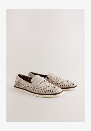 Pár béžových pletených kožených slip-on mokasín s bielymi podrážkami a tmavým lemom, vystavených na svetlom neutrálnom pozadí.
