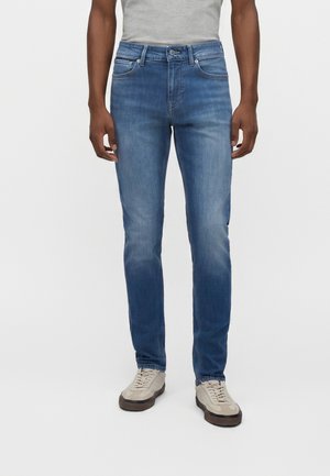 Homme portant un jean slim bleu moyen et des baskets gris clair, debout sur un fond blanc uni.
