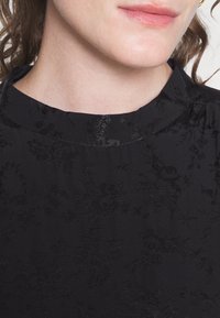 Black fabric with a floral pattern. The collar is rounded, and the texture is slightly raised, adding depth to the monochromatic design.