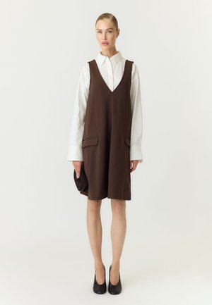 Donna che indossa una camicia bianca a maniche lunghe sotto un vestito marrone senza maniche, tiene in mano una pochette marrone abbinata e indossa tacchi neri.