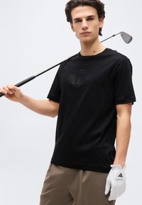 Νεαρός άνδρας που φοράει μαύρο πουκάμισο και μπεζ παντελόνι, κρατώντας ένα γκολφ κλαμπ στον ώμο του και φοράει λευκό γάντι γκολφ στο αριστερό χέρι.