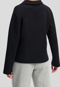 Sudadera negra con cuello, de corte holgado y mangas largas. Textura suave y dobladillo recto; combinada con pantalones de chándal grises.