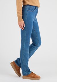 Jeans de mezclilla azul ajustados, con un diseño clásico de cinco bolsillos y una cintura de altura media. Llevados con zapatillas de ante marrón.