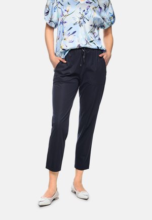 Woman wearing navy drawstring pants, blue floral short-sleeve blouse, and white pointed-toe flats, standing with hands in pockets.
