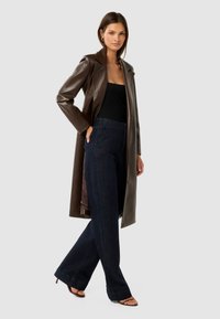 Hnedý kožený trench kabát cez čierny top, spárovaný s tmavomodrými širokými nohavicami a čiernymi lodičkami na podpätku. Jednoduchý, elegantný dizajn.