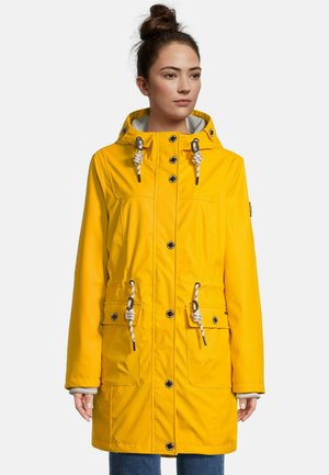 Frau trägt einen gelben Regenmantel mit Kapuze, Seilzügen, Druckknöpfen, Vordertaschen und grauem Innenfutter, stehend vor einem weißen Hintergrund.