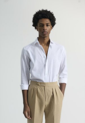 Camisa de lino blanca con cuello abotonado y mangas remangadas, combinada con pantalones de tela beige con pliegues y detalle de botón. Fondo neutro.