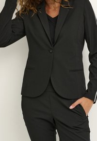 Blazer noir au design ajusté, fermeture à un bouton, revers crantés et poches latérales ; fabriqué en tissu lisse.