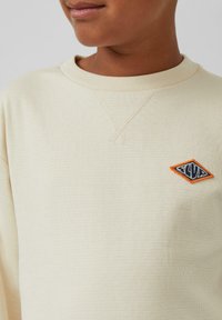 Lichtbeige lange mouwen shirt met ribbeltextuur. Heeft een oranje en marineblauw geborduurd diamant-logo aan de linkerkant.