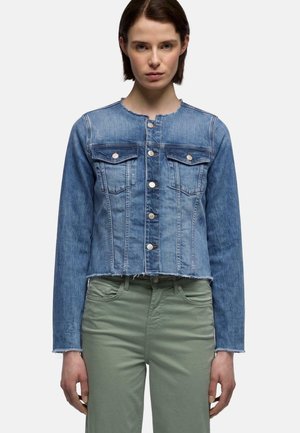 Vrouw draagt een blauwe spijkerjas met metalen knopen en groene hoge taille broek tegen een effen lichte achtergrond.