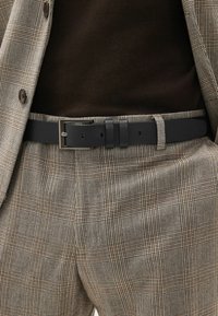 Ceinture en cuir noir avec une boucle en argent rectangulaire, portée sur un haut marron et un pantalon beige à motifs avec un design à carreaux subtil.