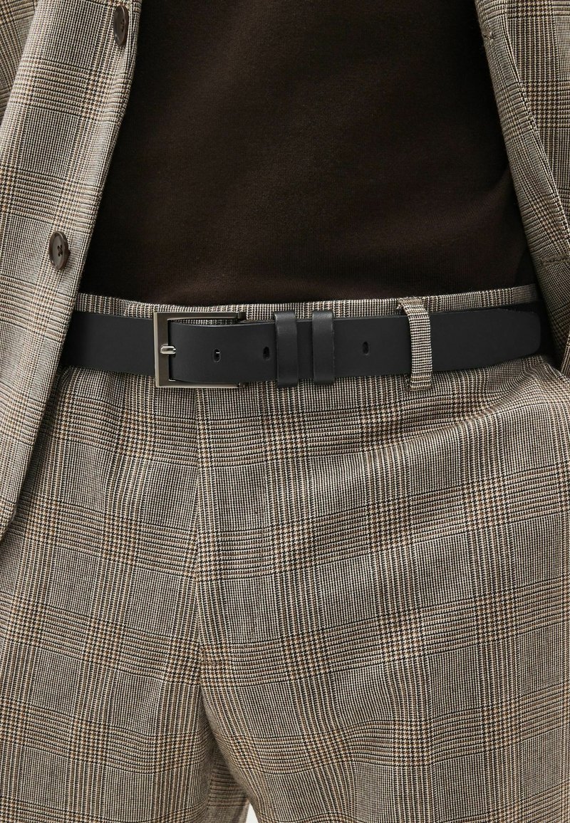 Schwarzer Leder Gürtel mit einer rechteckigen silbernen Schnalle, getragen über einem braunen Oberteil und gemusterten beigen Hosen mit einem subtilen Karodesign.