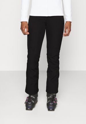 Person trägt schwarze Skihose und schwarze Skischuhe mit pinken und silbernen Schnallen, steht vor einem schlichten weißen Hintergrund.