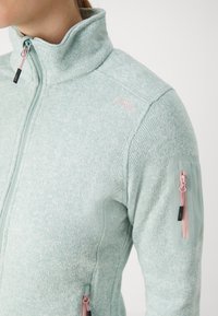 Femme portant une veste en maille vert clair avec des tirettes de fermeture éclair roses et une petite poche zippée sur la manche supérieure.