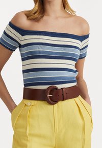 Top sin mangas a rayas en azul y crema, combinado con pantalones amarillos de pliegues y un cinturón marrón con una hebilla redonda trenzada.