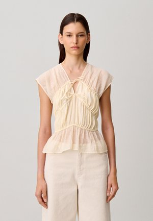 Jeune femme aux longs cheveux foncés portant un chemisier beige texturé sans manches avec bas à volant et un pantalon crème taille haute sur fond uni.